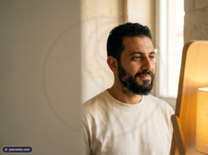 رجل في منتصف الثلاثينيات بلحية داكنة يتأمّل بهدوء نحو مرآة دافئة في غرفة كريميّة، يفكّر في تساقط شعر اللحية